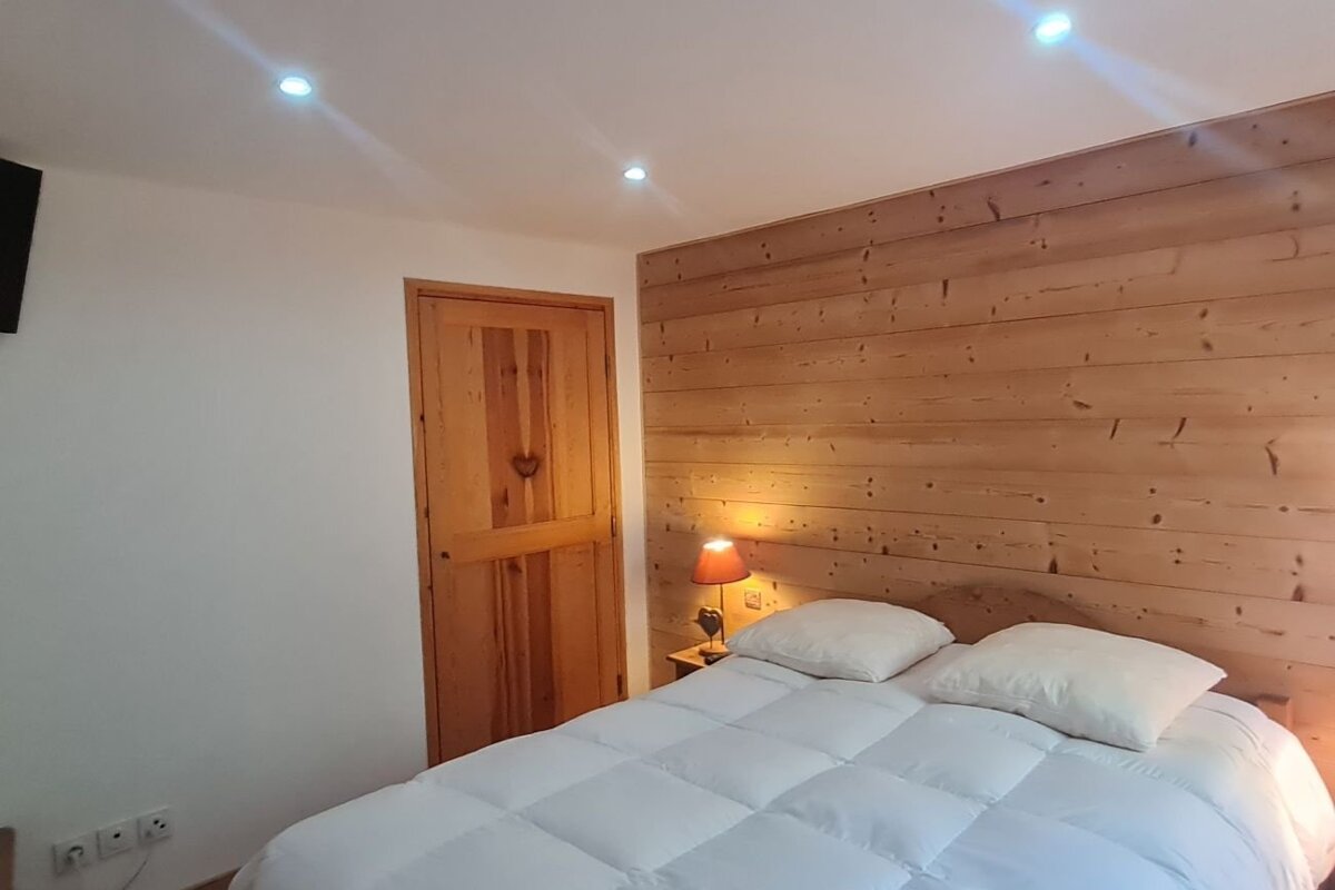 Cozy bedroom featuring a bed with a white duvet, a rustic wood-paneled wall, matching wooden door, and a glowing bedside lamp.
