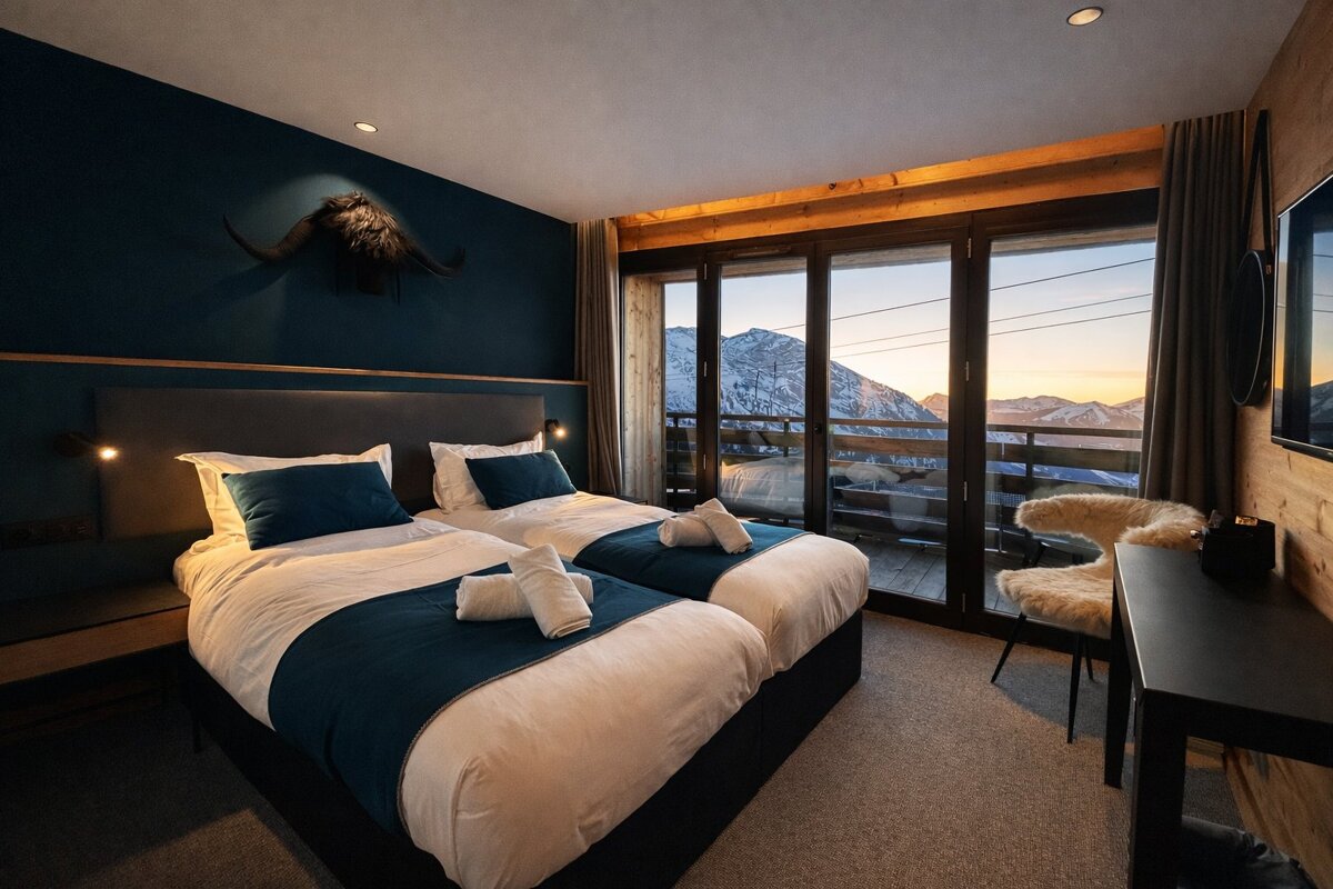 A cozy bedroom with two twin beds and large glass doors revealing a scenic view of snowy mountains at sunset.