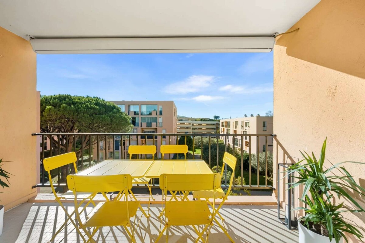 A sunny balcony featuring bright yellow outdoor table and chairs, a potted plant, and a view of apartment buildings, green trees, and a clear blue sky.