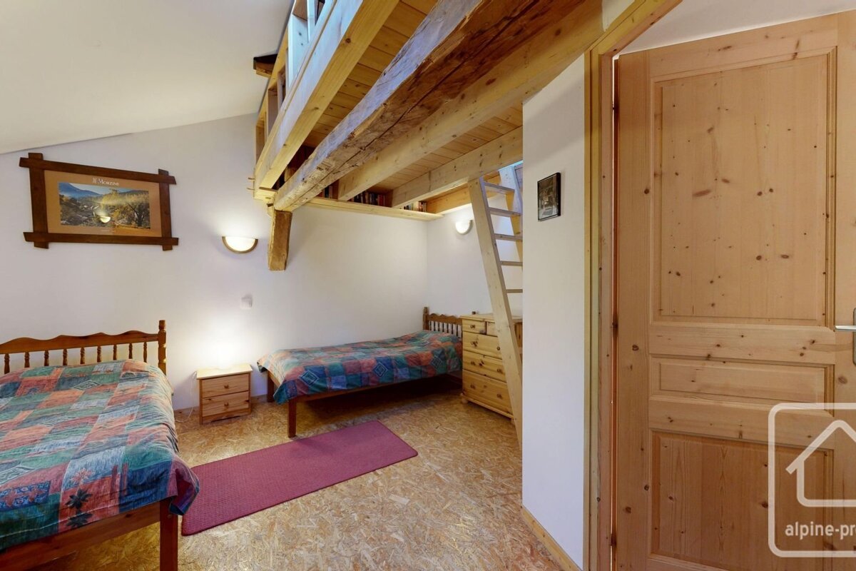 Rustic bedroom with two single beds, a wooden loft above accessed by a ladder, white walls, and wood-paneled door.