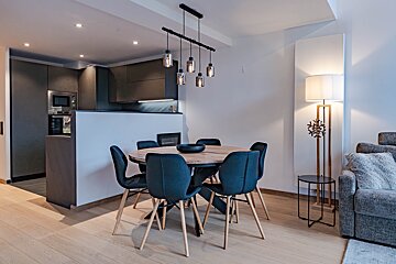 Modern open-plan kitchen, dining area with a round wooden table and blue chairs, and a cozy living space with a grey sofa and floor lamp.