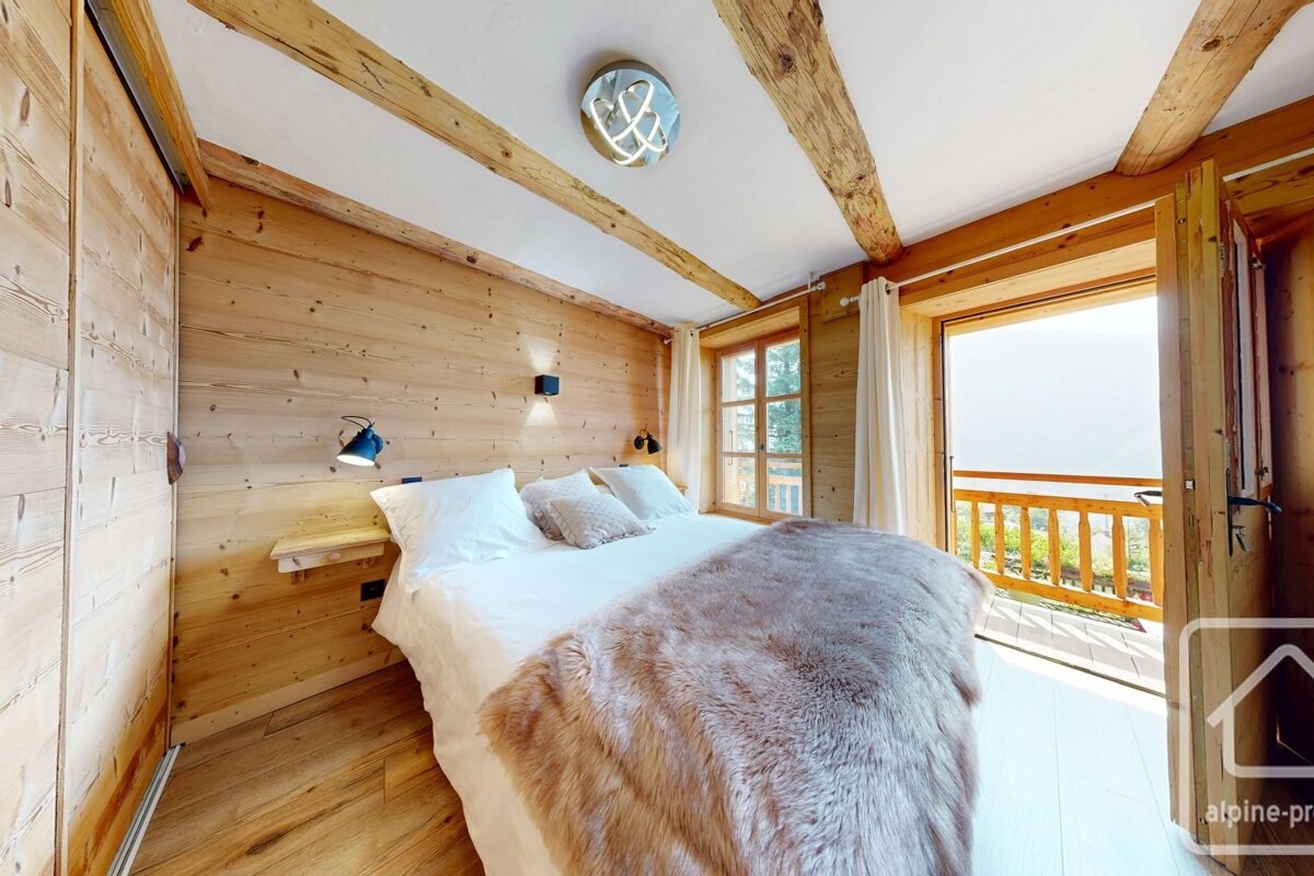 A cozy, wood-paneled bedroom features a large bed with a fur throw and a balcony overlooking a scenic mountain view, bathed in natural light.