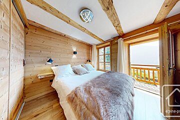 A cozy, wood-paneled bedroom features a large bed with a fur throw and a balcony overlooking a scenic mountain view, bathed in natural light.