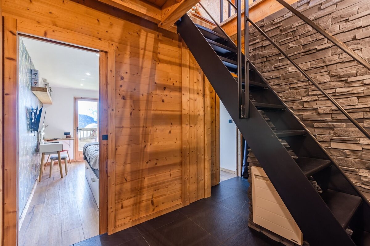 Rustic modern cabin interior featuring a black metal staircase next to a stone wall, with a doorway opening into a bedroom.