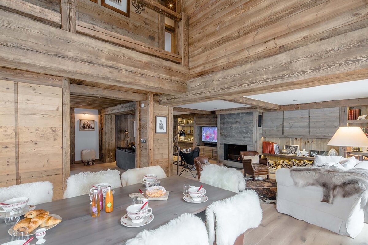 A rustic wooden chalet interior with a dining table set for breakfast, and a cozy living room featuring a fireplace, TV, and comfortable seating.