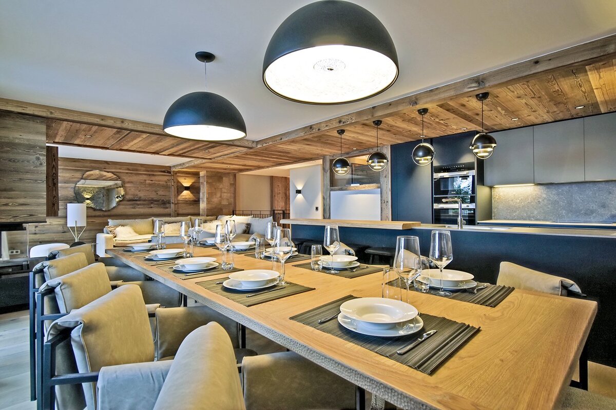 A stylish rustic-modern dining room with a long table set for guests, an open-plan kitchen, and warm wooden paneling throughout.