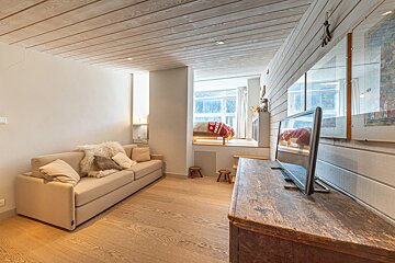 Cozy wooden cabin-style studio featuring a beige sofa, rustic TV stand, and an elevated sleeping nook in the background.