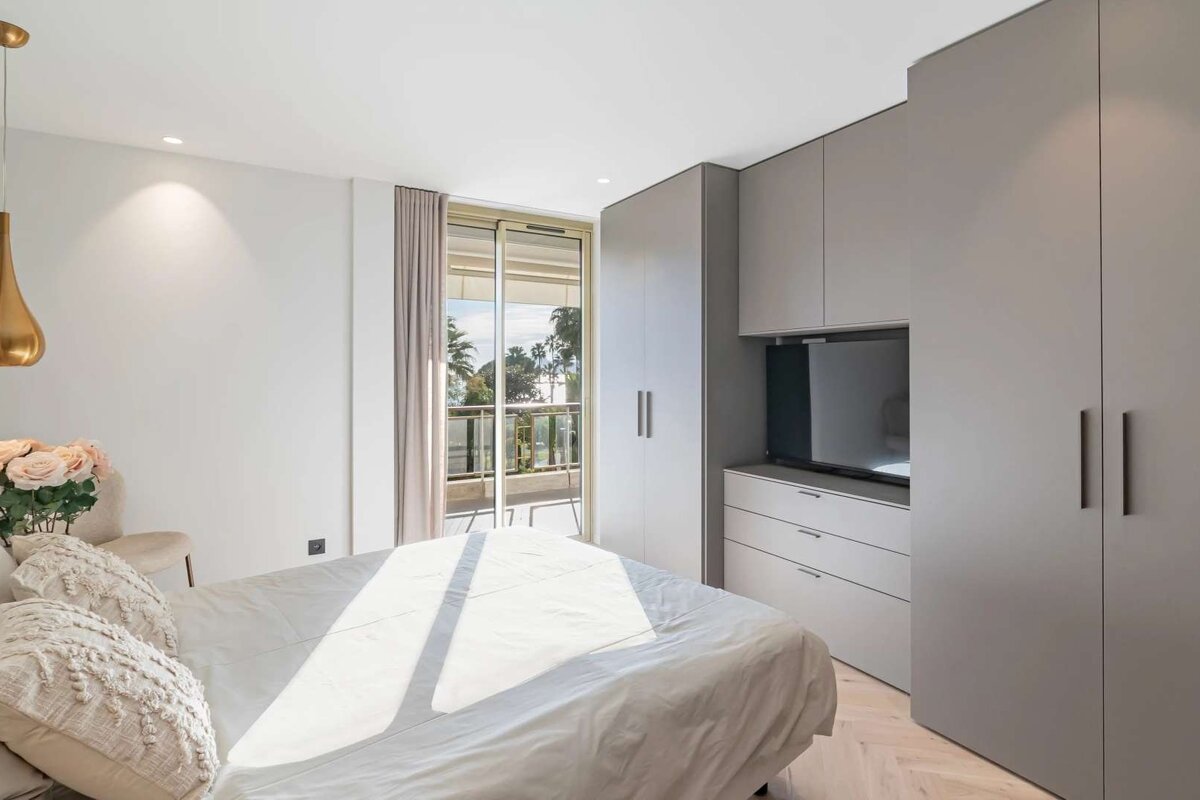 A bright, modern bedroom with a white bed, sleek gray built-in wardrobes and TV unit, and a balcony offering a view of palm trees.