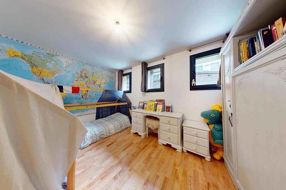 A bright child's room with a huge world map mural, bunk bed, white desk, and wooden floor. A stuffed dinosaur peeks out from a wardrobe.