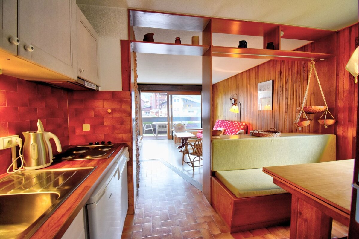 Cozy cabin-style apartment featuring a wooden kitchen, dining nook with a green bench, and living area leading to a balcony.