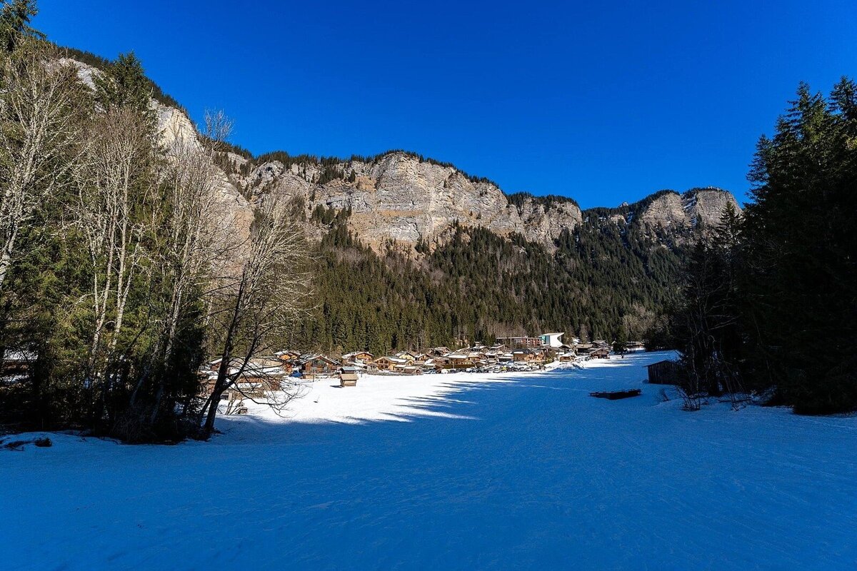 A snowy valley with a charming village nestled against towering, forested mountains under a brilliant blue sky on a clear winter day.