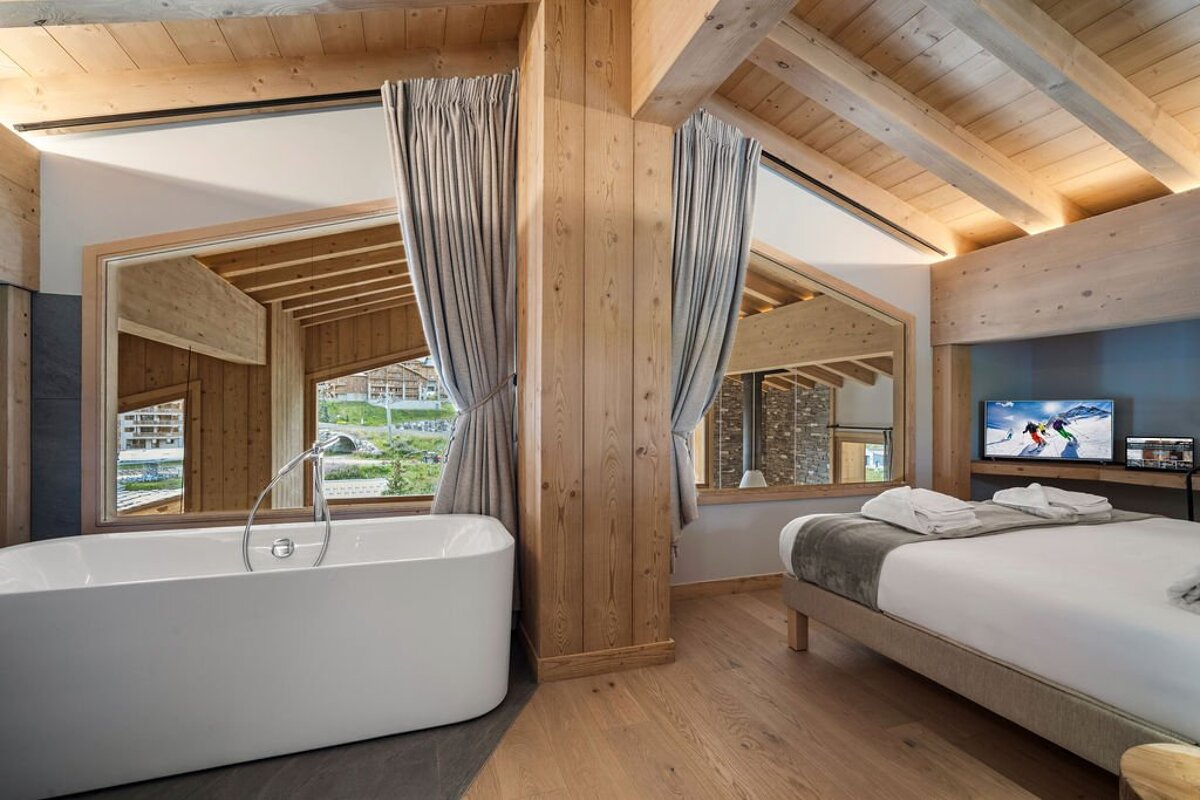 Luxurious wooden hotel suite with a freestanding tub, comfy bed, a TV, and panoramic window views of green mountains.