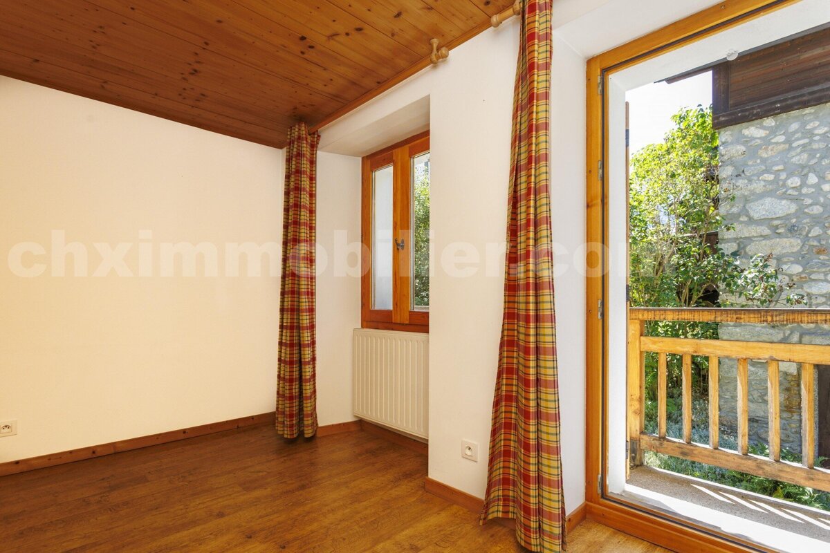 An empty room with wood floors/ceiling, white walls, and plaid curtains framing windows/balcony door. A balcony overlooks trees and a stone building.