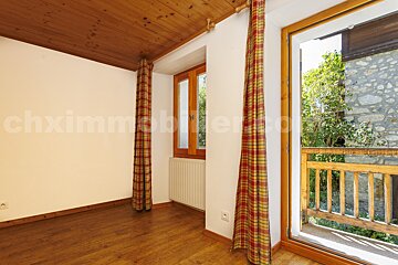 An empty room with wood floors/ceiling, white walls, and plaid curtains framing windows/balcony door. A balcony overlooks trees and a stone building.
