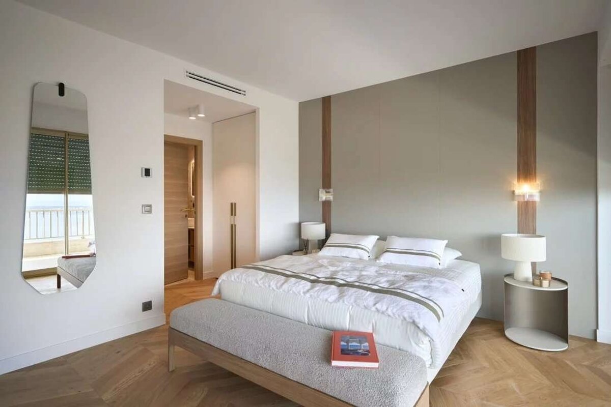 A stylish, modern bedroom features a white bed, grey accent wall with wood panels, herringbone wood floor, and a mirror reflecting a balcony view.