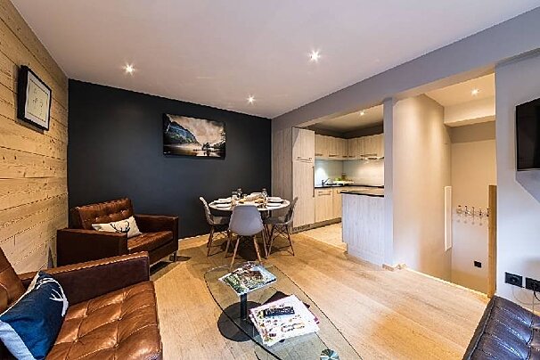 Modern apartment interior featuring brown leather armchairs, a glass coffee table, a dining set, and a compact kitchen.