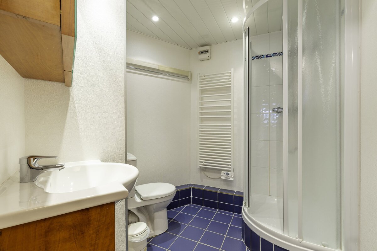 A compact bathroom with a white sink, toilet, corner shower, and blue tiled floor. Features a wood vanity and a white towel warmer.