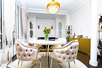 Bright, elegant dining space featuring a white round table, tufted beige chairs, a gold chandelier, and fresh roses. Golden accents throughout.