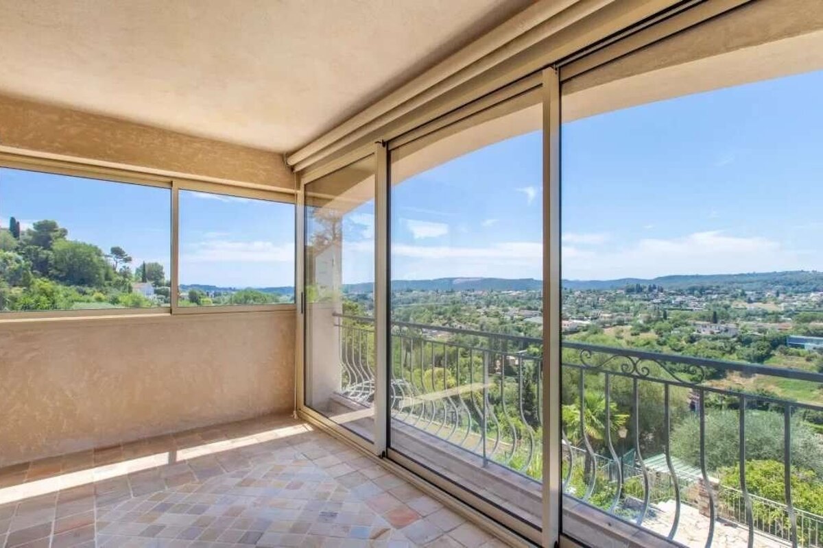 Bright balcony with large glass doors and windows, offering a panoramic view of a green valley, distant town, and blue sky.