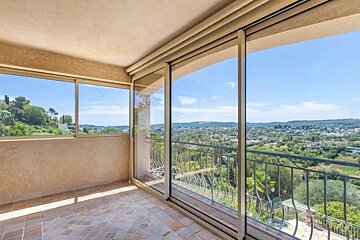 A sunny balcony with a tiled floor and glass doors offering a panoramic view of a lush green valley and distant town under a bright blue sky.