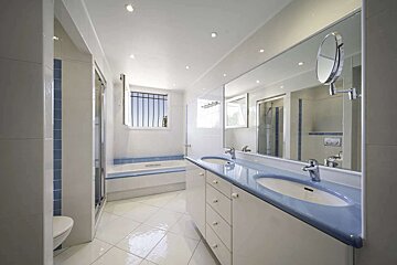 A modern white and blue bathroom featuring a double vanity, large mirror, bathtub, shower, toilet, and window with security bars.