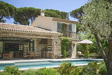 A beautiful Mediterranean-style villa with a terracotta roof, stone accents, outdoor dining, and a pool, surrounded by lush trees under a clear sky.