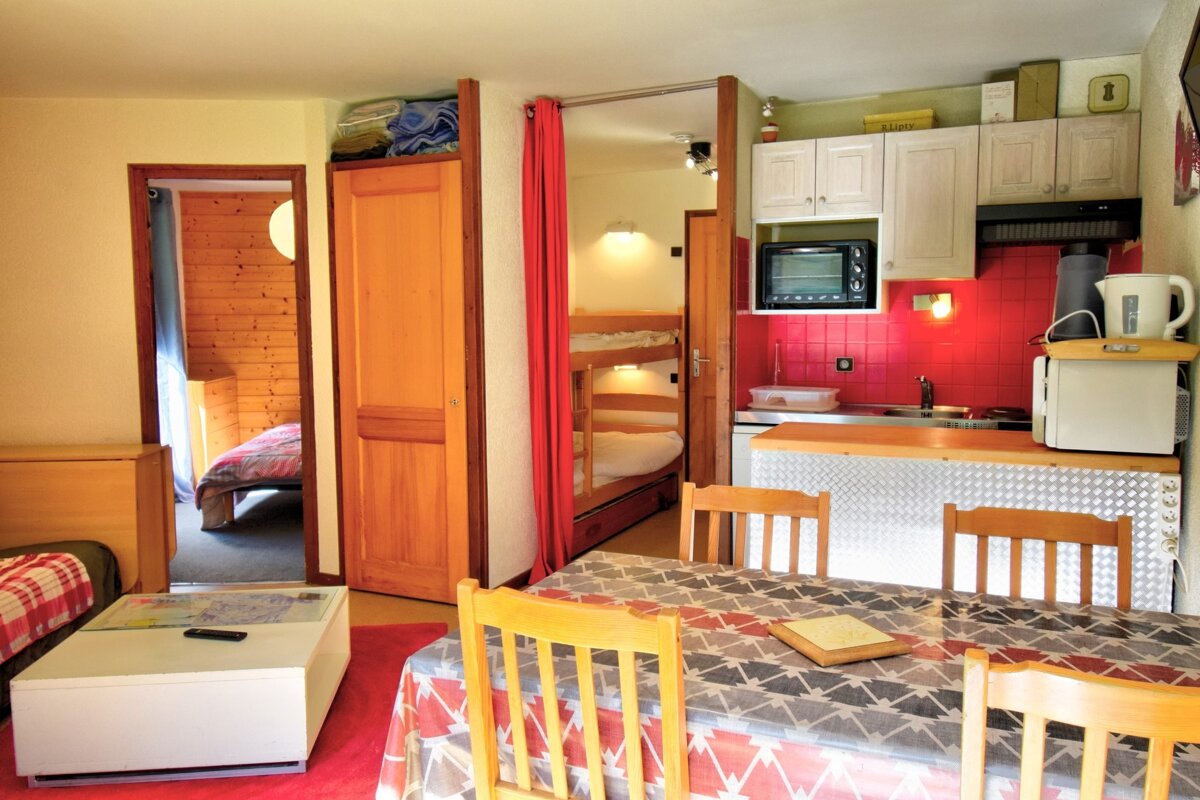 A compact living space with a red-tiled kitchenette, dining table, and doors leading to a bedroom and a bunk bed area.