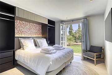 A chic bedroom with a large white bed, dark built-in closets, an armchair, and open doors leading to a bright garden.