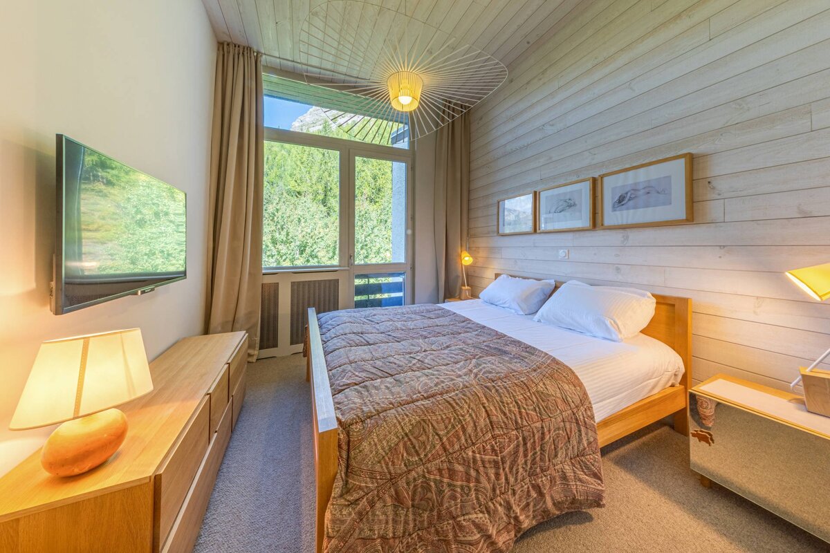 A cozy wood-paneled bedroom featuring a double bed, unique wire pendant light, and large glass doors looking out onto trees.