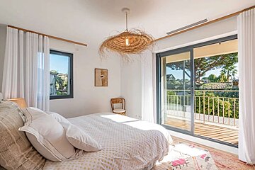 A bright bedroom with a large bed, unique straw pendant light, and sliding glass doors to a sunny balcony overlooking lush greenery.