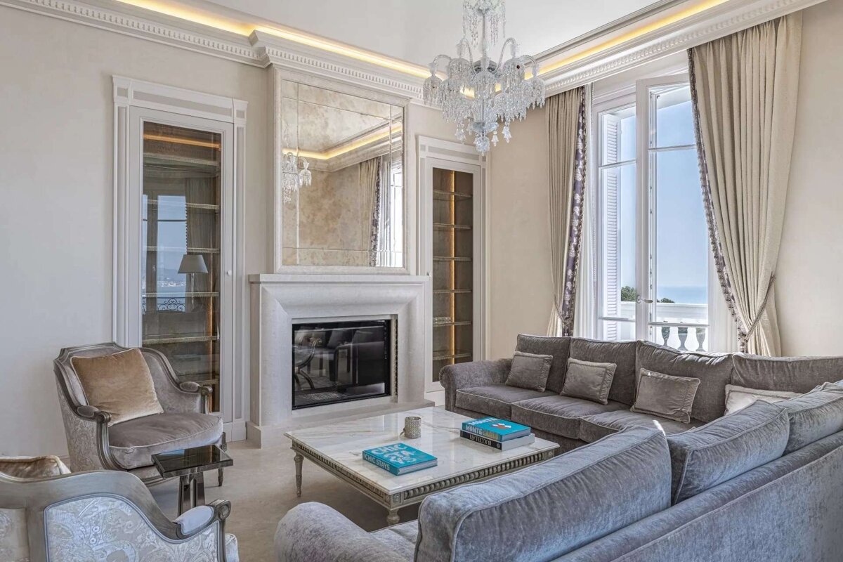 An opulent living room with plush grey seating, a grand fireplace with mirror, dazzling chandelier, and large windows offering a beautiful sea view. Elegant decor.