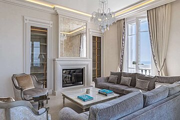 An opulent living room with plush grey seating, a grand fireplace with mirror, dazzling chandelier, and large windows offering a beautiful sea view. Elegant decor.