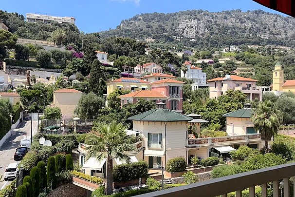 A charming, colorful hillside village with houses and lush trees, backed by a large mountain under a clear blue sky.