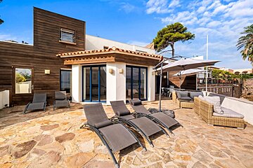 Sunny modern villa with wood and white walls, a stone patio featuring lounge chairs, outdoor seating, and umbrellas under a clear blue sky.