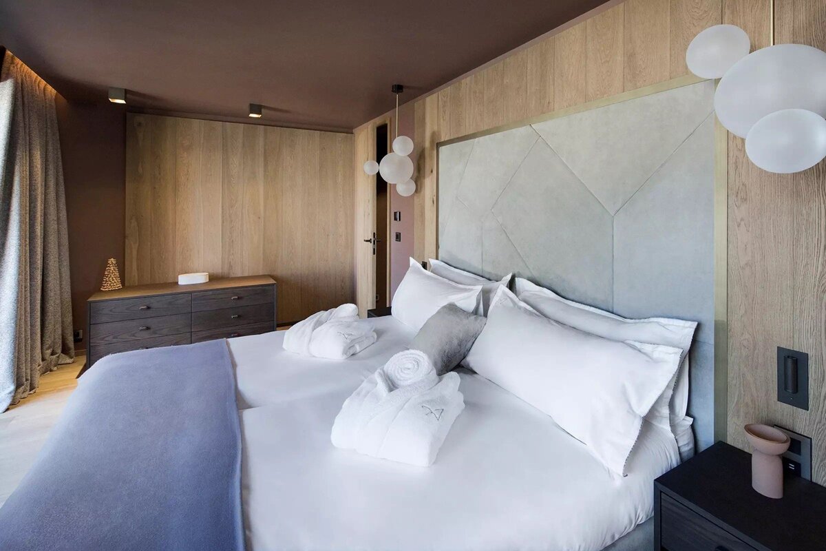 A modern hotel bedroom with a large bed, white linens, folded bathrobes, a gray geometric headboard, and warm wood paneled walls.