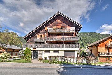 A large multi-story wooden chalet with balconies, situated in a scenic mountain area under a cloudy blue sky.
