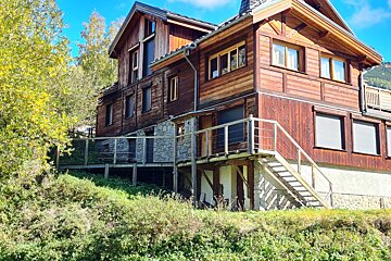 A large, multi-story wooden and stone chalet with decks and stairs, nestled on a sunny, green hillside under a bright blue sky.