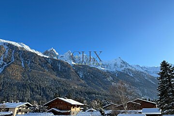 Snow-covered chalets in a valley with majestic, pine-covered mountains and snowy peaks under a clear blue sky. CHX watermark._