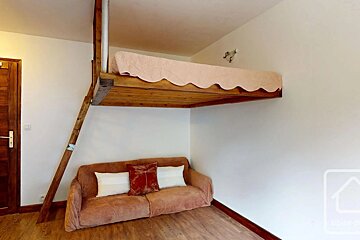 A wooden loft bed with a pink scalloped blanket is suspended over a cozy brown sofa, accessible via a wooden ladder in a minimalist room.