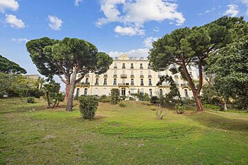 An elegant, cream-colored building stands on a grassy hill, framed by two large pine trees, under a blue sky with white clouds.
