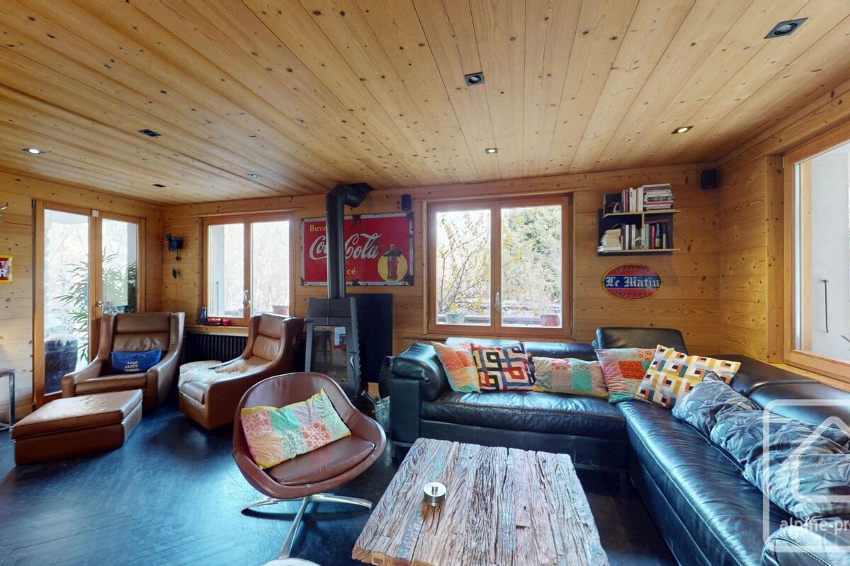 A cozy chalet living room with wood-paneled walls/ceiling, black leather sofa, brown armchairs, a wood stove, rustic coffee table, and large windows.