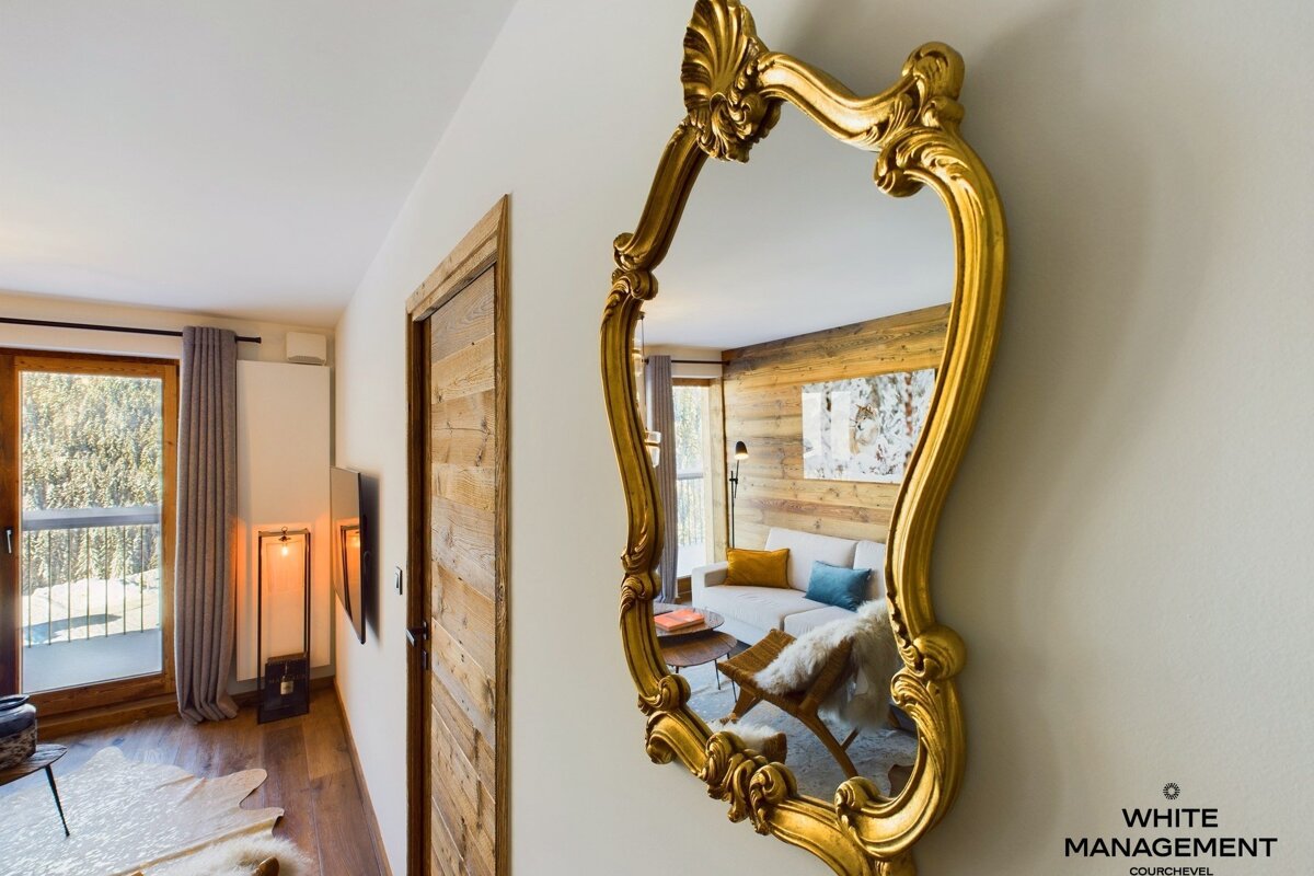 An ornate gold mirror reflects a cozy living room with wood paneling and a white sofa, while a balcony overlooks a forest.