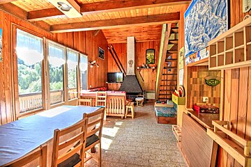 Rustic wooden cabin interior with a dining table, a living area with a fireplace, and large windows overlooking a forest.
