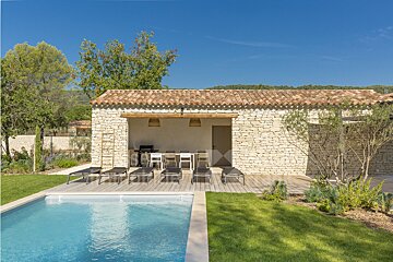 A beautiful stone villa with a terracotta roof, outdoor dining, loungers, and a BBQ on a wooden deck, beside a clear blue swimming pool and green lawn.