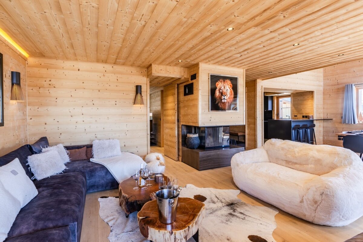 A cozy wooden chalet living room with a blue sectional, fluffy white sofa, log coffee tables, and a fireplace featuring a lion portrait.
