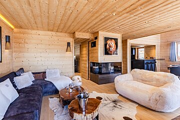 A cozy wooden chalet living room with a blue sectional, fluffy white sofa, log coffee tables, and a fireplace featuring a lion portrait.