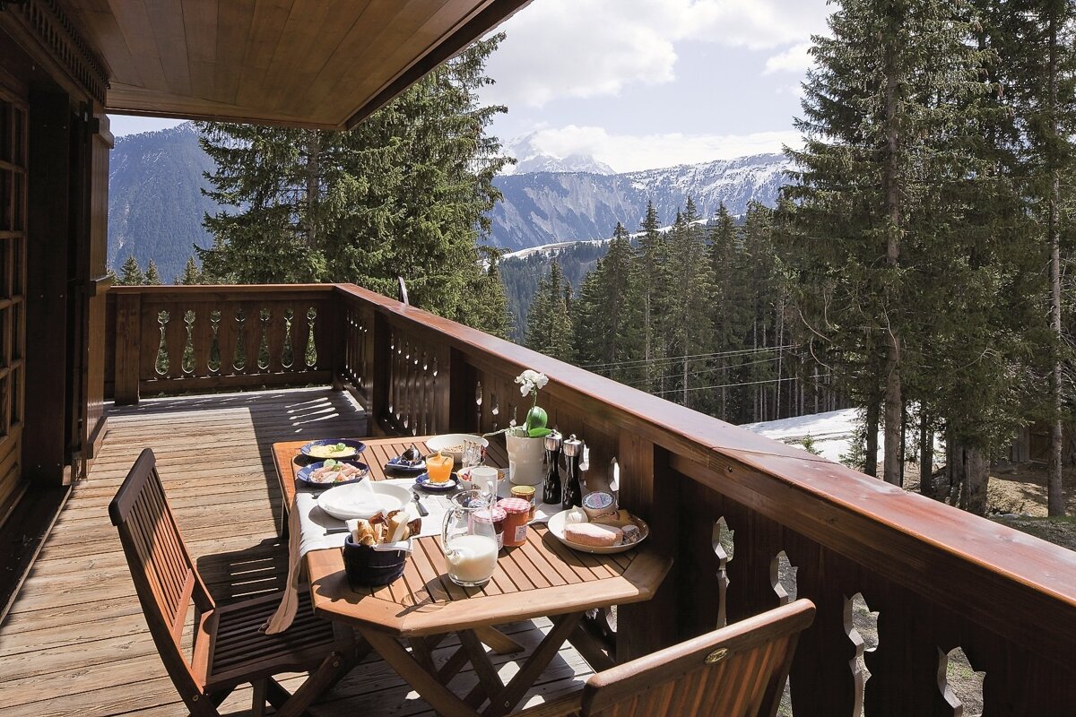 A sunlit wooden balcony with a breakfast spread overlooks snowy mountains and a dense pine forest.