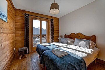 Cozy, rustic bedroom with wood-paneled walls, two beds, and a balcony with mountain views. Sunlight fills the comfortable space.