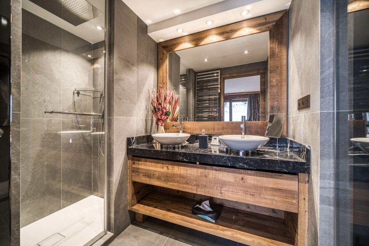 A modern bathroom features a glass shower, gray tiled walls, and a rustic wooden double vanity with a black marble countertop and vessel sinks.