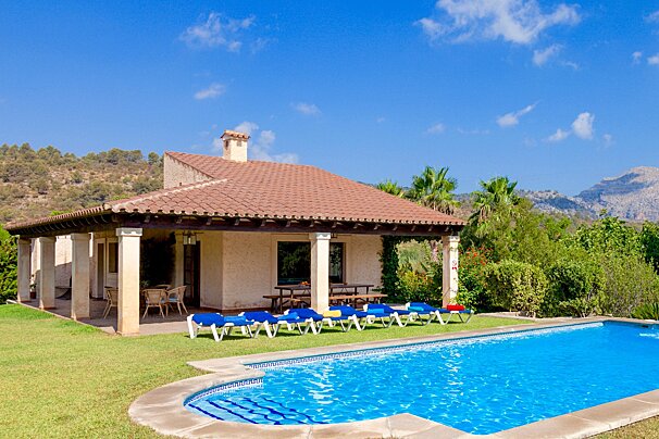 A charming Mediterranean villa with a red-tiled roof, a sparkling blue swimming pool, and a green lawn, nestled against mountains under a clear blue sky.